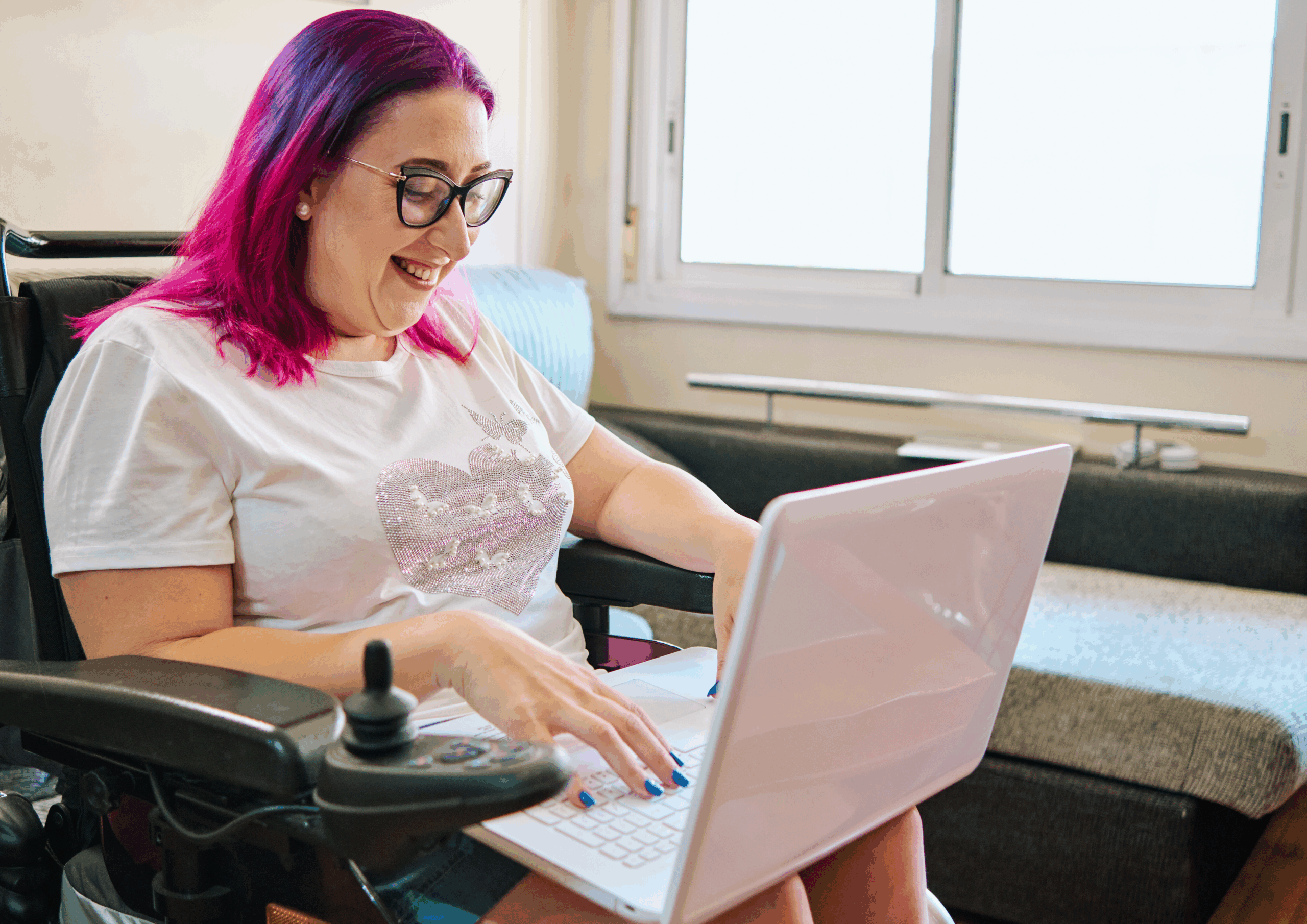 Lady with pink hair sitting in wheelchair, working on a laptop on her lap.